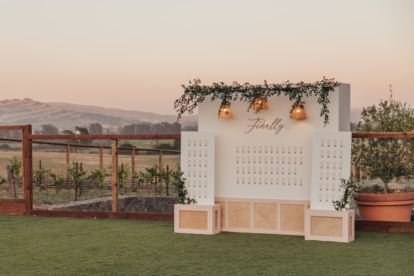 A wedding seating chart display adorned with greenery and hanging lights, set against a scenic landscape at sunset.