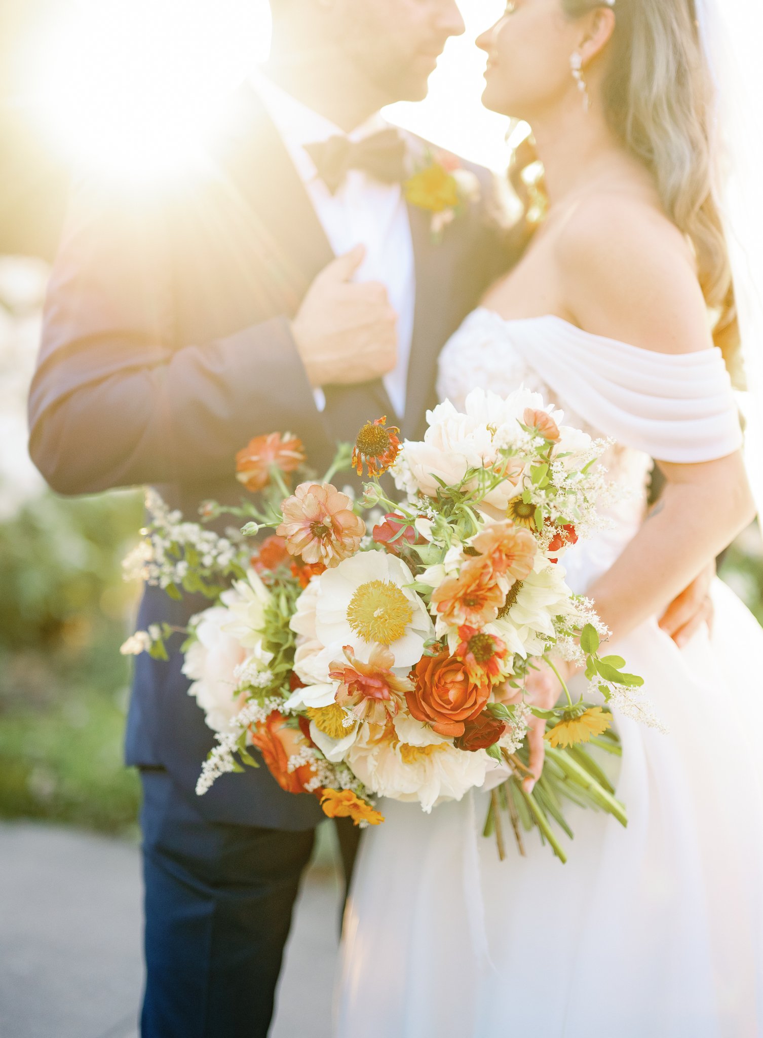 Bride and groom standing close with the sun shining behind them, holding a bouquet of white and orange flowers.