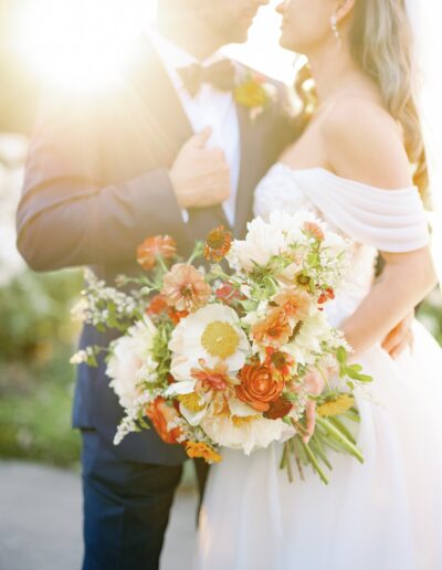 A bride and groom stand close together outdoors in the sunlight, with the bride holding a large bouquet of white and orange flowers.