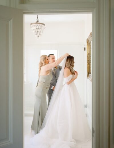 A woman adjusts another woman's veil while a man stands beside them in a sunlit room. They are all dressed formally, with the woman in white wearing a wedding dress.