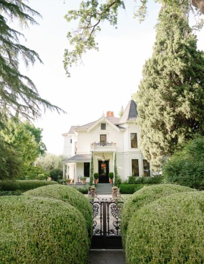 Victorian-style house with a pointed turret, surrounded by lush greenery and a wrought-iron gate in front.