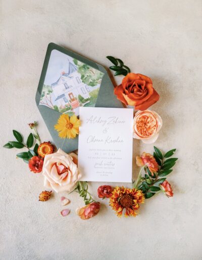 A wedding invitation card surrounded by various flowers and an envelope featuring a house illustration.