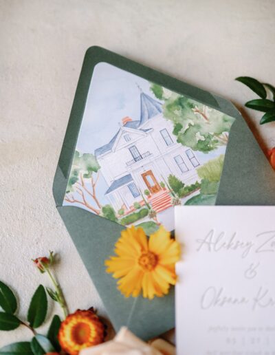 A green envelope with a watercolor illustration of a house is accompanied by a bouquet of orange flowers and a partially visible invitation card.