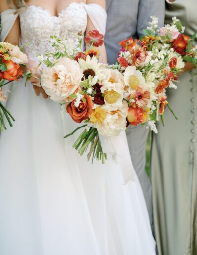 Three people holding bouquets of mixed flowers, including roses and peonies, with two in light-colored attire, one in a silver-gray suit.