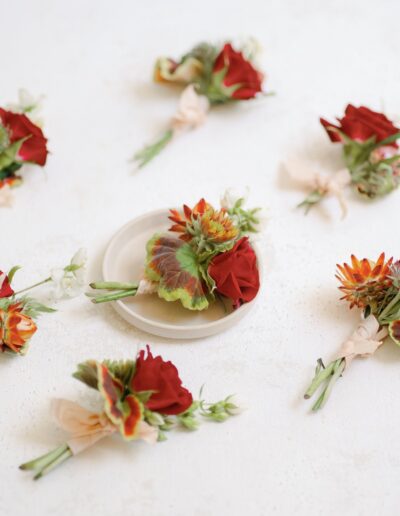 Several small floral arrangements with red roses and green leaves scattered on a white surface, with one placed on a small white plate in the center.