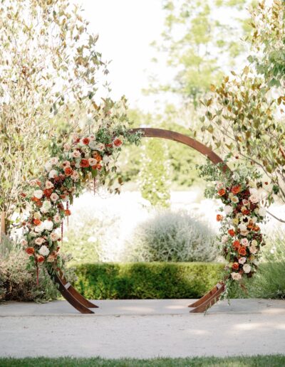 A circular wooden arch decorated with red, orange, and white flowers stands outdoors amid greenery.