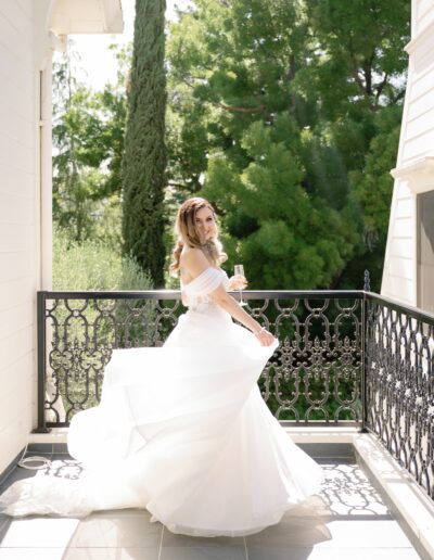 Bride in a white wedding dress holding a champagne glass stands on a sunlit balcony with trees in the background.