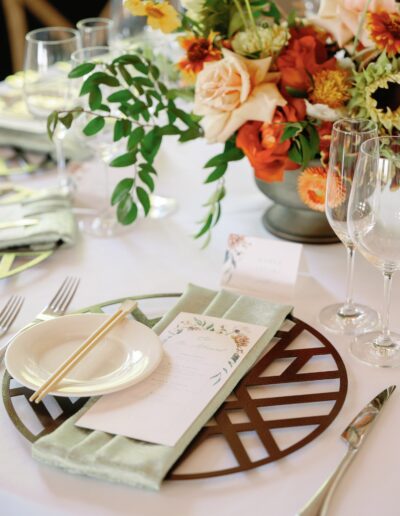 Elegant table setting with a floral centerpiece, a menu placed on a green napkin, a small plate with chopsticks, and glassware on a white tablecloth.