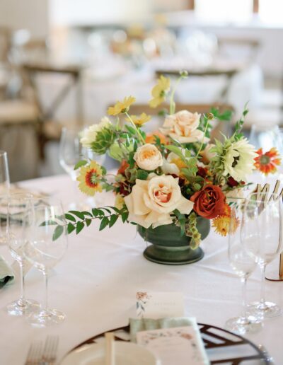 Table setting with floral centerpiece featuring cream and orange flowers, greenery, wine glasses, and a menu on a white tablecloth.