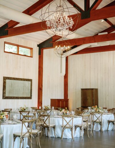 An elegant dining hall with wooden beams, round tables with white tablecloths, floral centerpieces, and a chandelier. Large mirror on the wall and a cabinet in the corner.