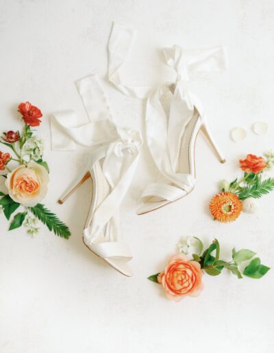 White high-heeled shoes with ribbon ties surrounded by orange and white flowers on a light background.