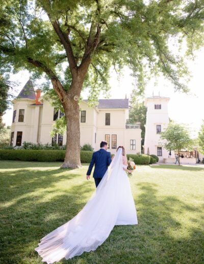 A bride and groom walk across a sunlit lawn towards a large, elegant house, with the bride's veil flowing behind them.