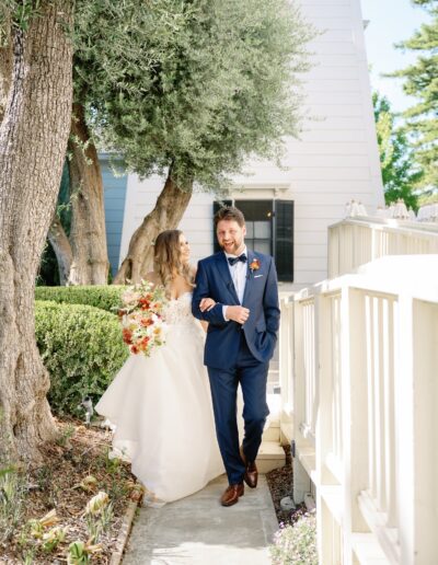 A bride and groom walk joyfully along a garden path. The bride holds a bouquet of flowers, and the groom wears a blue suit. Trees and a white building are in the background.