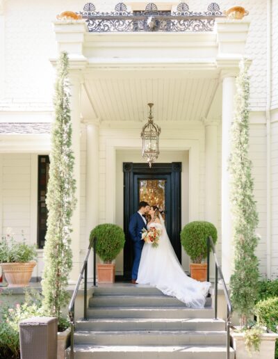 A bride and groom share a kiss on the steps of an elegant white building with black double doors, flanked by topiary plants and tall greenery.