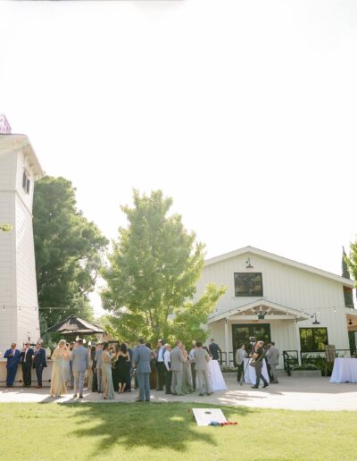 A group of people gathered outside a barn-style building with a tower on a sunny day. Trees and lawn surround the area.