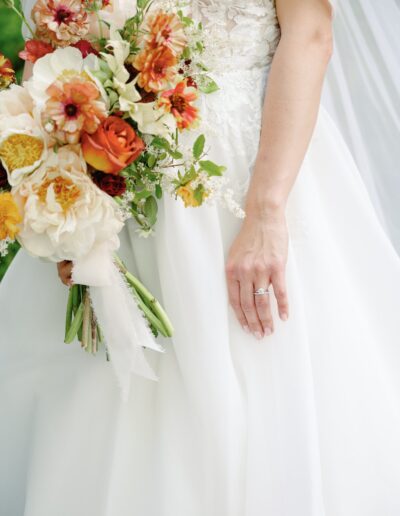 Bride in white dress holds a bouquet of orange, white, and yellow flowers. Her left hand shows a silver ring.