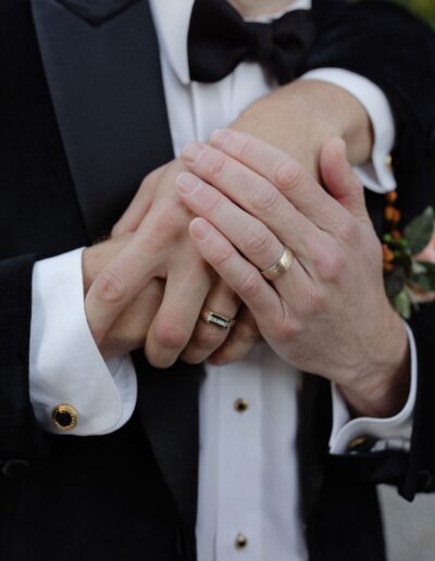 Two people wearing rings clasp hands. Both are in formal attire, including a suit jacket and dress shirt with cufflinks.