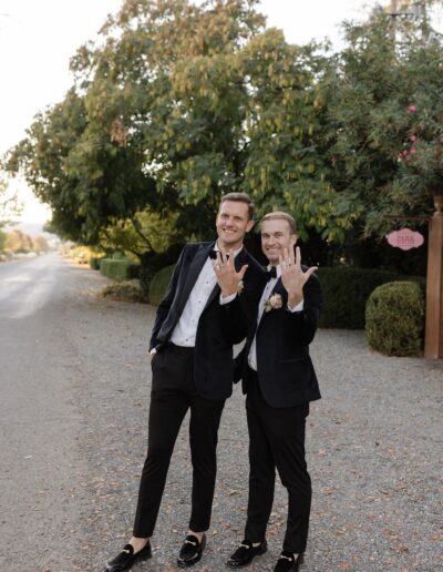 Two men in formal attire stand on a path, smiling and showing their rings. Trees and bushes line the path in the background.