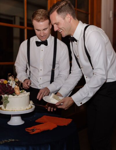 Two people in formal attire serve slices from a decorated cake on a stand over a dark blue tablecloth.