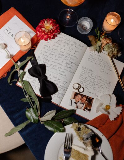 Handwritten letters, a bow tie, flowers, candles, and wedding rings on a table with cake and drinks.