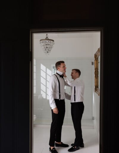 Two men in formal attire stand in a well-lit room with tiled walls. One adjusts the other's bow tie. A chandelier hangs overhead.