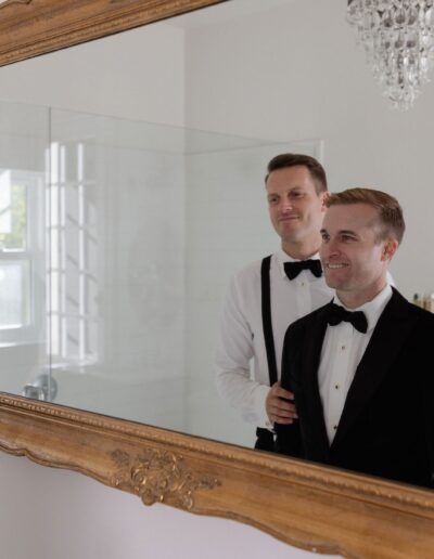 Two men in formal attire stand in front of a large ornate mirror. One adjusts the other's bow tie. A chandelier and a sink are visible in the background.