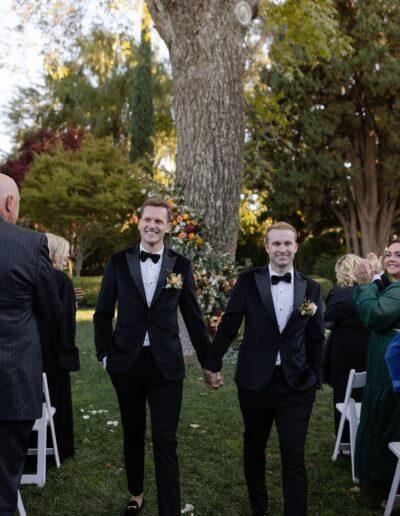 Two men in tuxedos hold hands and smile, walking outdoors in a garden ceremony with trees and seated guests around them.