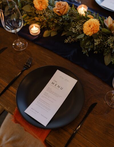 A black plate with a menu sits on a wooden table. Nearby are a black fork and knife, lit candles, and an arrangement of orange flowers and greenery.