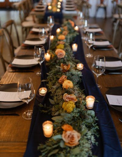 A wooden table is set for a formal meal with plates, glasses, and utensils. A centerpiece of candles, greenery, and orange flowers runs along a dark table runner.