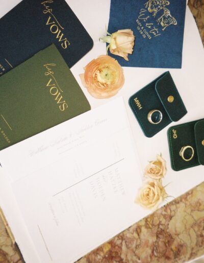 Wedding invitation, vow booklets, two rings in green cases, and beige roses on a marble surface.