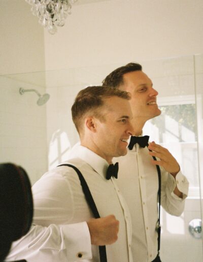 Two men in white dress shirts and bow ties stand in front of a mirror adjusting their outfits, smiling. They appear to be getting ready for a formal event.