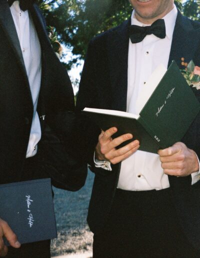 Two men in suits holding green booklets, standing outdoors. One man has a floral boutonniere.