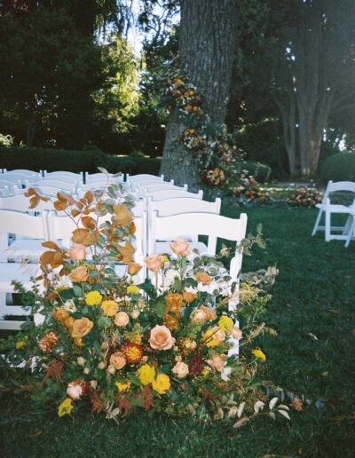 White folding chairs arranged outdoors with lush floral decorations of orange and yellow flowers on a grassy area, surrounded by tall trees.