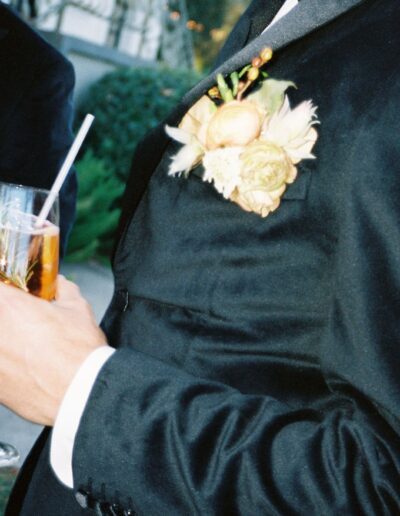 Two people in formal black attire hold drinks, each with a floral boutonniere. One person is holding a glass with a straw.