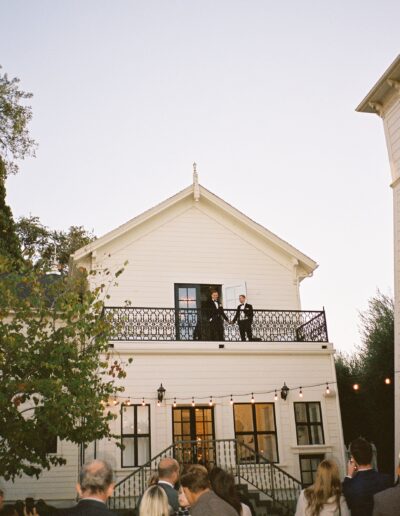 A crowd of people stands in front of a two-story house with a balcony, where two people are speaking to the gathered audience.