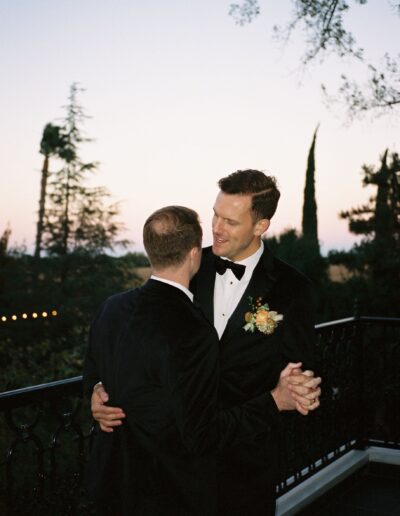 Two people in tuxedos dance closely on an outdoor balcony at dusk, with trees in the background.