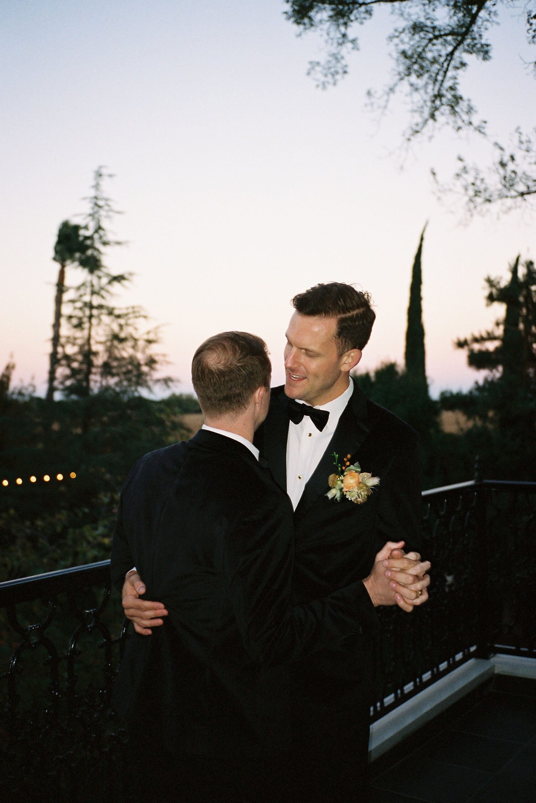 Two people in tuxedos dance closely on an outdoor balcony at dusk, with trees in the background.