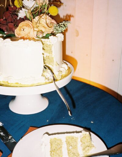 A sliced cake with white icing and floral decoration sits on a stand, next to a plate with a piece of the cake, on a blue tablecloth.