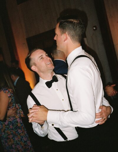 Two people in formal attire dance together, both wearing white shirts and dark suspenders. They are surrounded by other people in a dimly lit room.