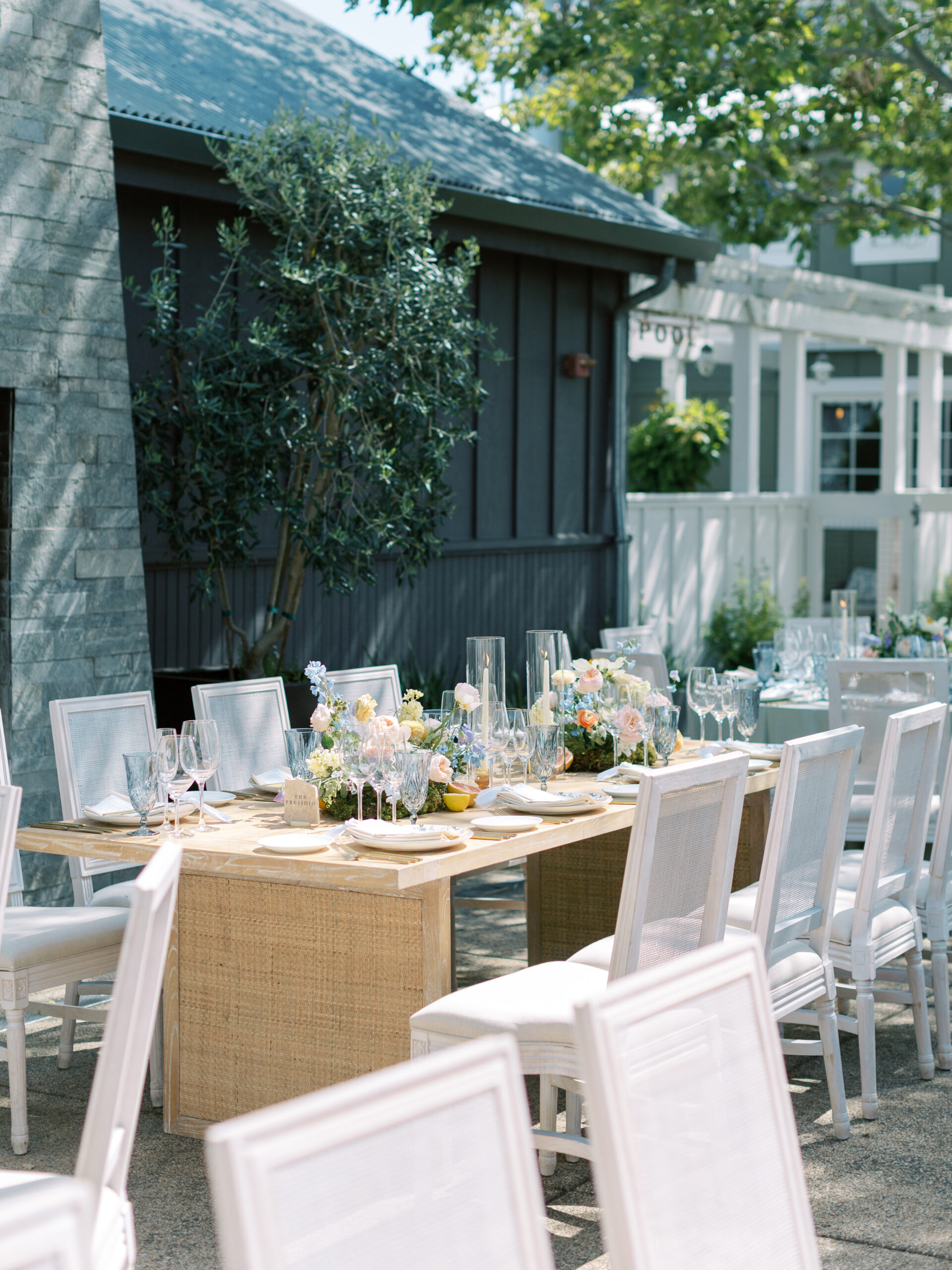 Outdoor dining area with a wooden table set with glassware, floral arrangements, and surrounded by white chairs. A building and trees are visible in the background.