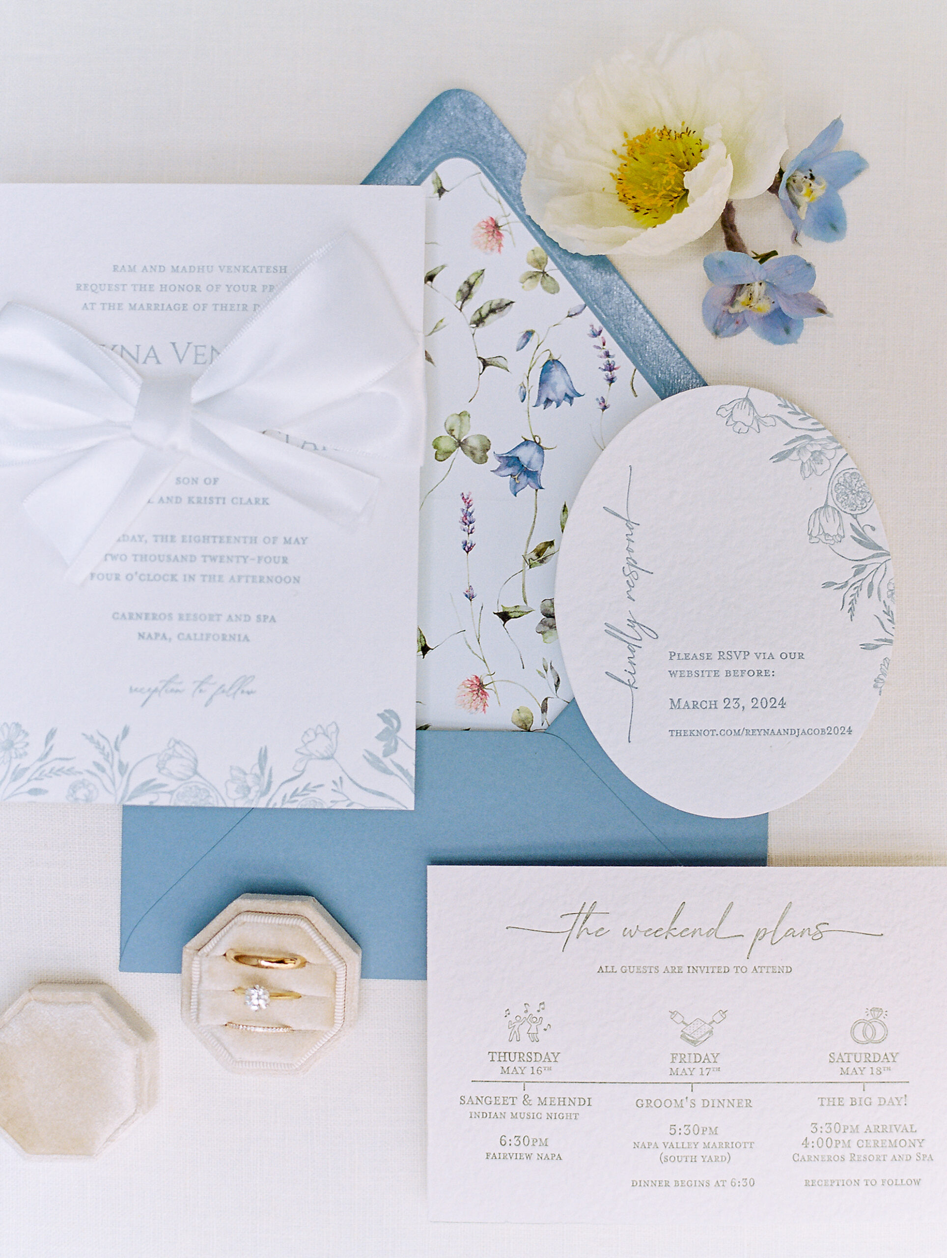 A flat lay of wedding invitations and cards with floral designs, a blue envelope, a white ribbon, a flower, and a hexagonal ring box with rings inside.