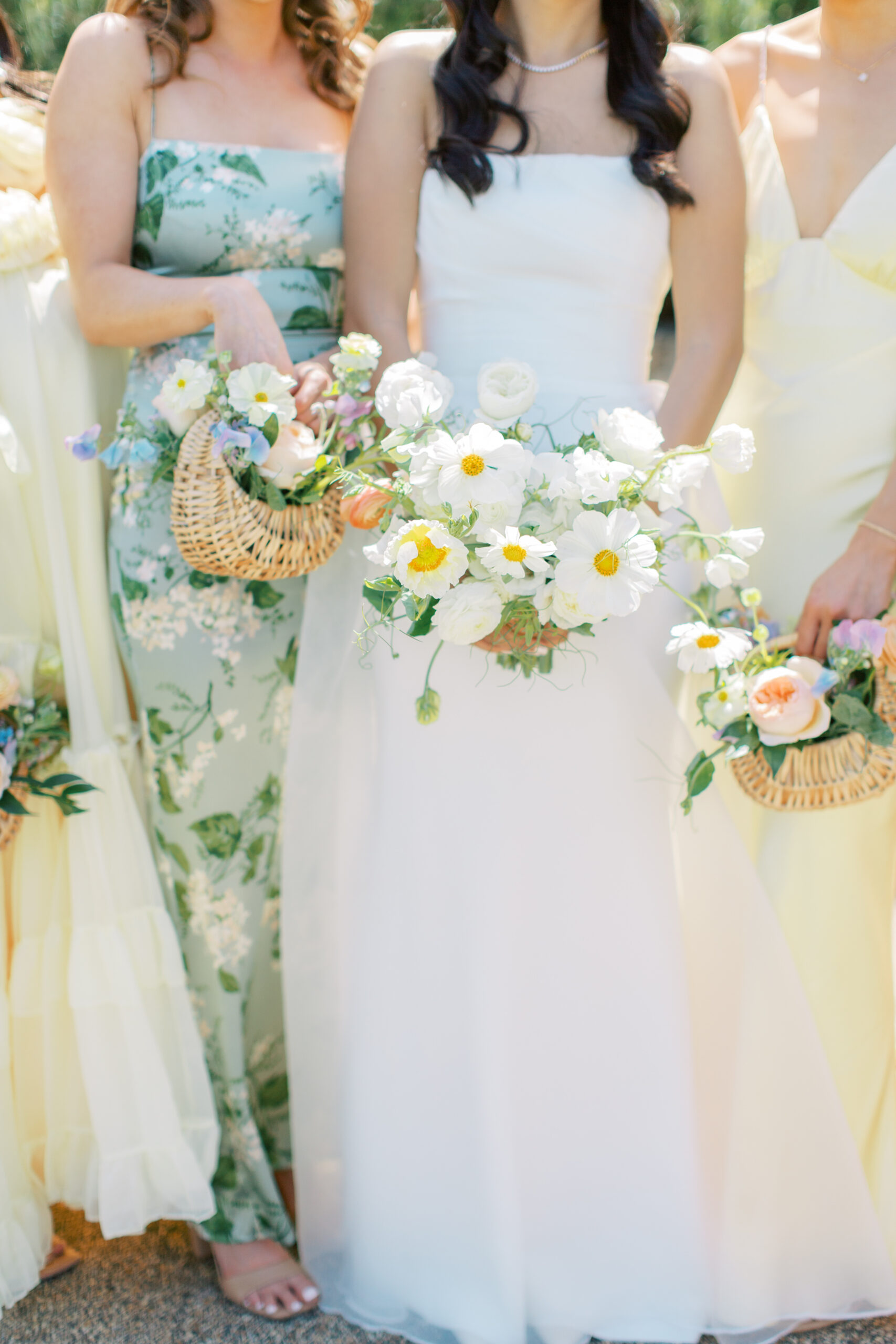 A bride in a white dress holds a bouquet of white and yellow flowers, surrounded by bridesmaids in pastel dresses also holding flowers.