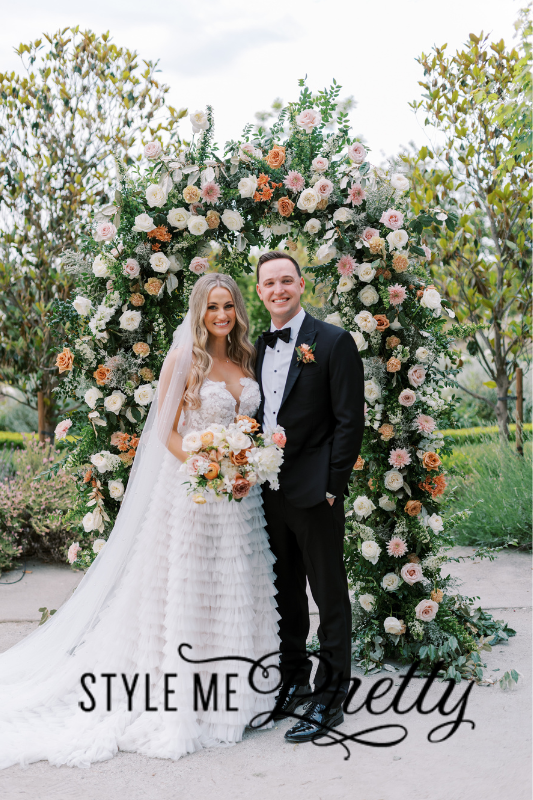 A bride and groom stand together in front of a floral arch outdoors, beautifully arranged by Tahoe wedding planners. She holds a bouquet, and they both smile happily at the camera.