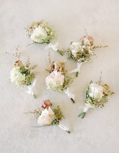 Six small floral boutonnieres with white and pink flowers and greenery arranged on a light textured background.