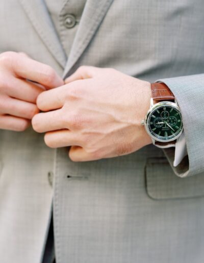 A person in a light gray suit adjusts their jacket. They wear a watch with a leather strap on their left wrist.