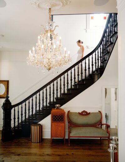 A person in a white outfit descends a dark wooden staircase in an elegantly decorated room with a chandelier and vintage furniture.