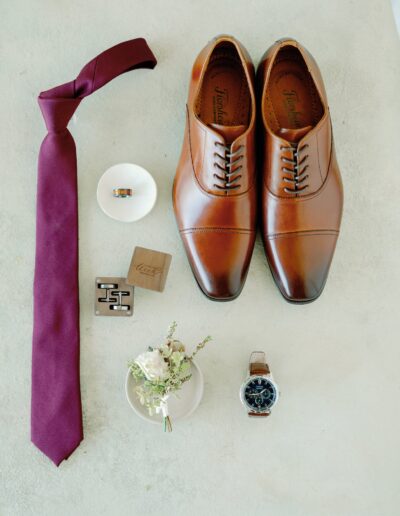 Brown leather dress shoes, a burgundy tie, cufflinks, two rings in small dishes, a wristwatch, a wooden box, and a small floral arrangement on a light surface.