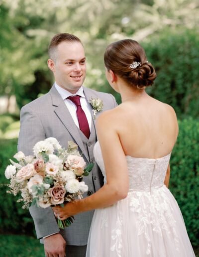 A bride and groom stand facing each other outdoors. The bride holds a bouquet of flowers and wears a strapless white dress. The groom wears a gray suit with a burgundy tie.