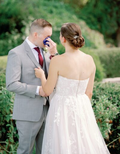 A bride in a white dress stands with a groom in a gray suit who is wiping his face with a blue cloth, surrounded by greenery.