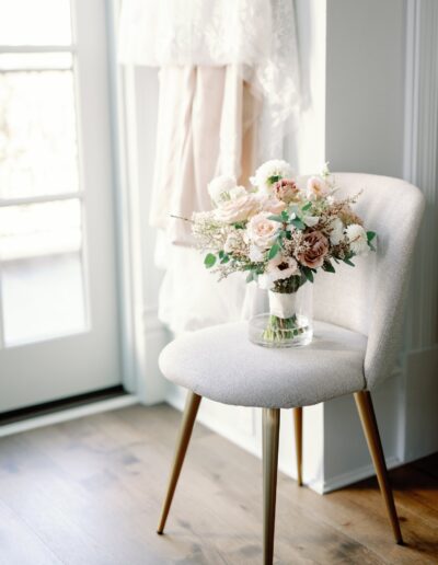 A vase of pastel flowers sits on a gray chair in front of a window. A white curtain and a light-colored dress are visible in the background.